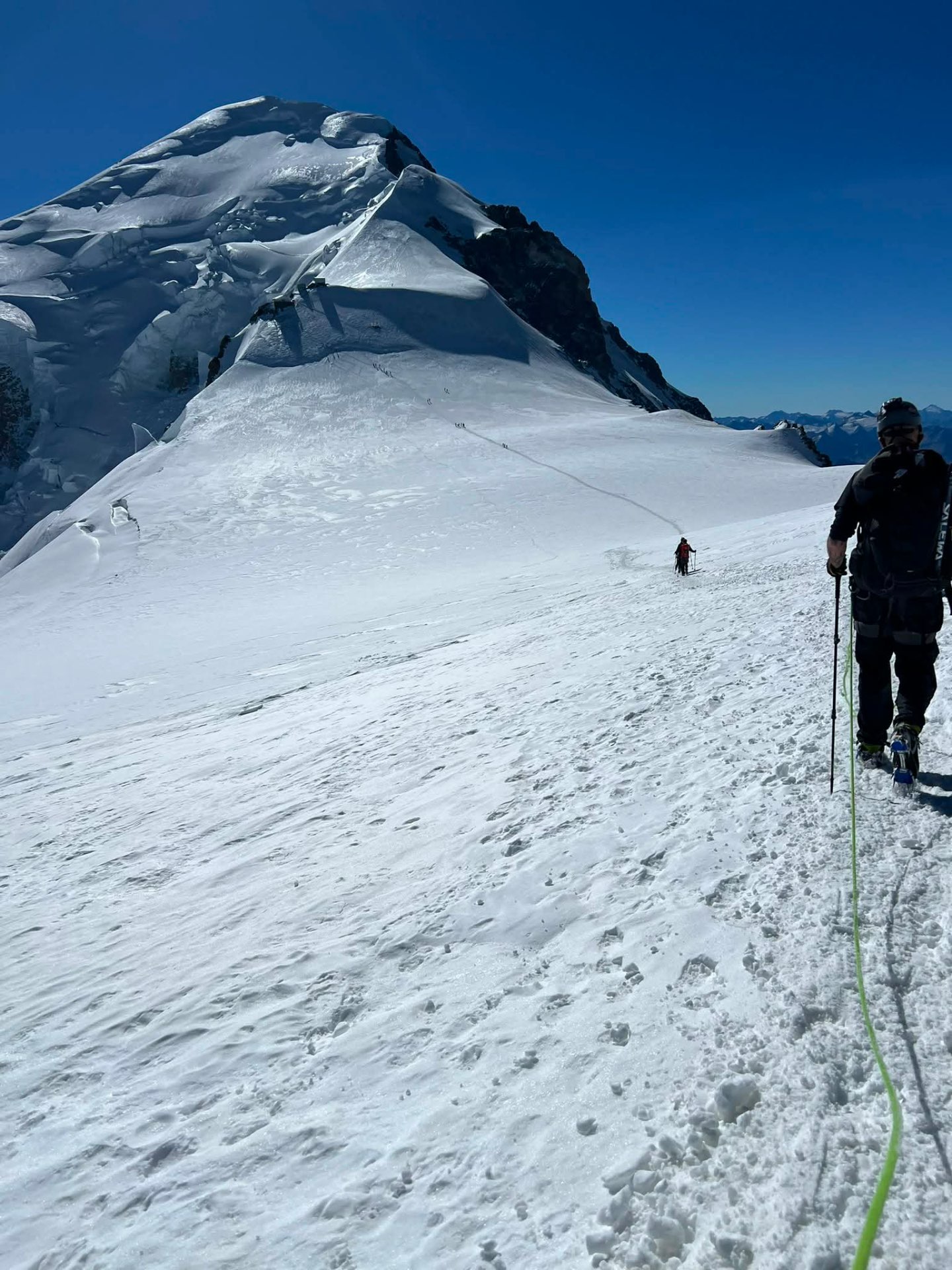 A roped climber walking a broad snow ridge with the summit dome ahead.